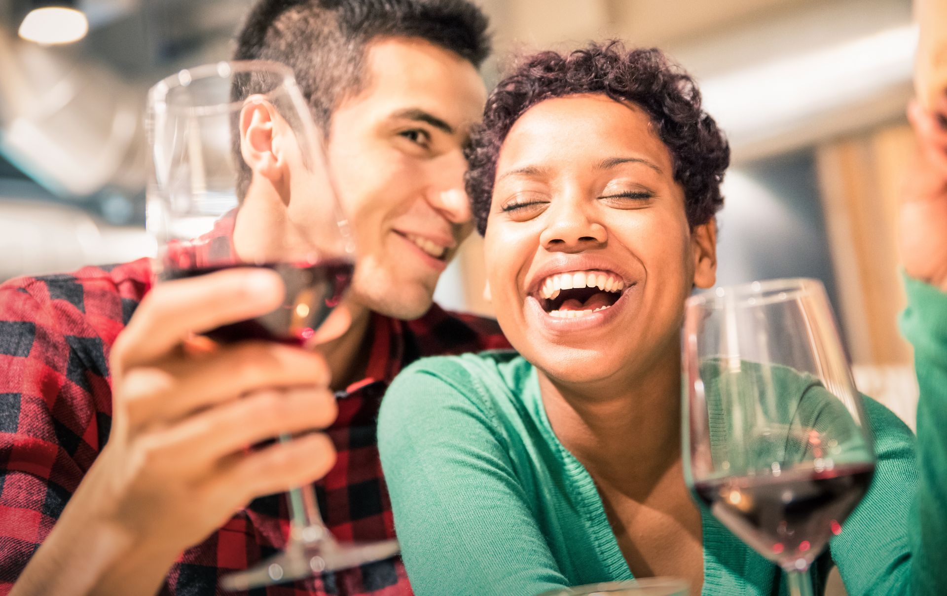 A joyful couple sharing a moment over glasses of red wine. The woman laughs wholeheartedly while the man leans in with a smile, creating a warm and lively atmosphere. The setting is cozy and inviting, perfect for an evening of connection and celebration.