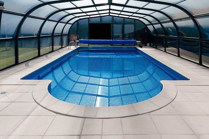 Wide view of an indoor swimming pool beneath a transparent enclosure with tiled deck and calm blue water.