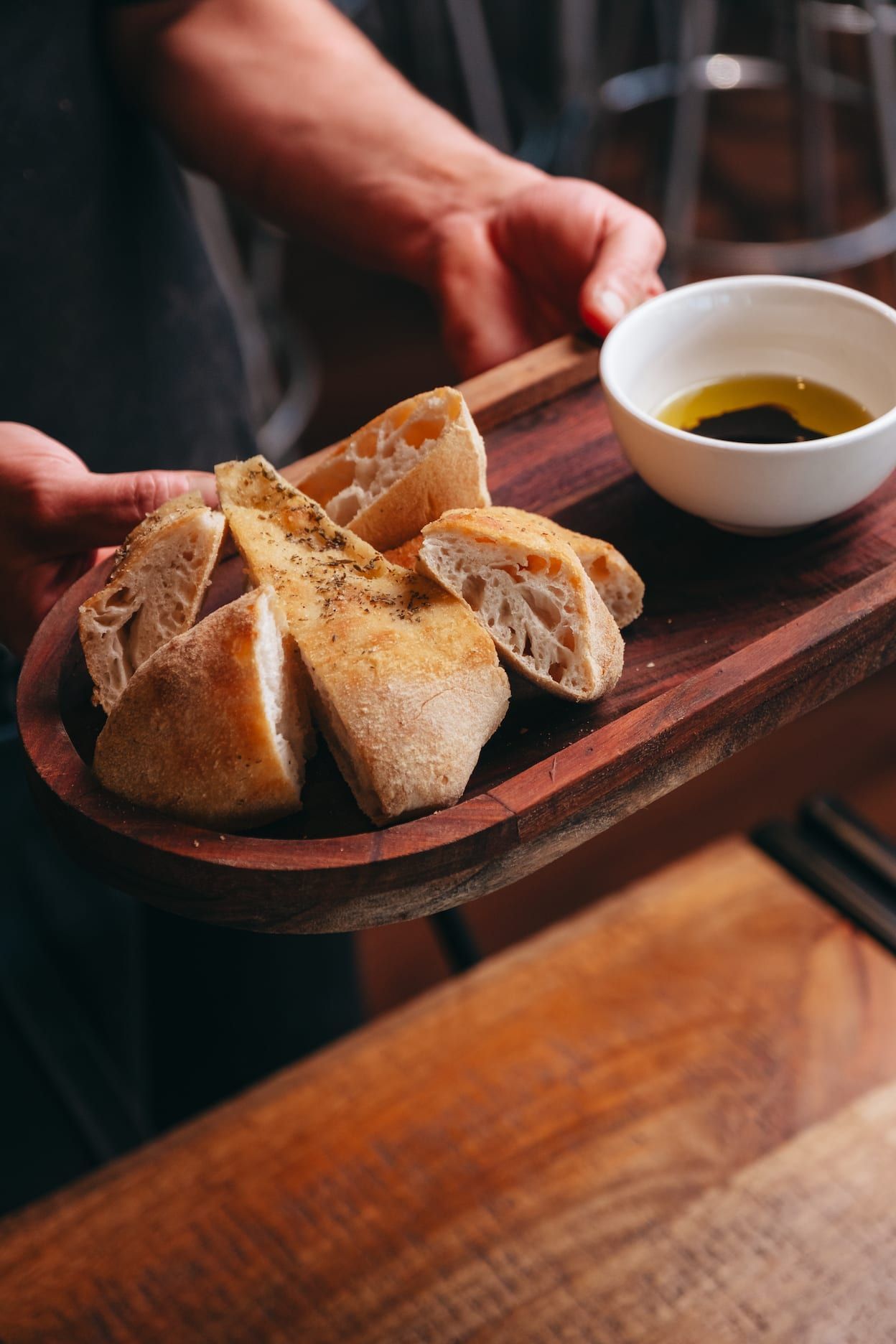 A person is holding a wooden tray of bread and a bowl of oil.