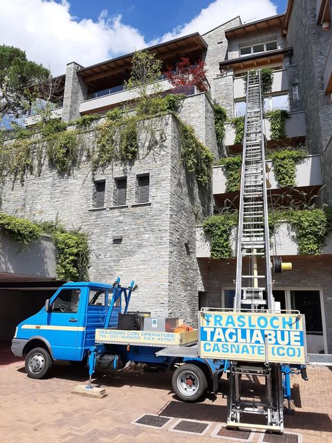 Camion blu con un cartello di traslochi parcheggiato accanto a un edificio ricoperto di rampicanti, sotto un cielo luminoso.
