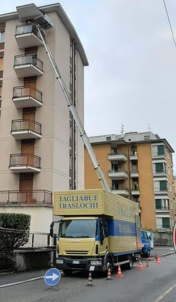 Grande camion giallo per traslochi parcheggiato accanto a edifici residenziali, con una gru che solleva oggetti fino a un balcone ai piani superiori.
