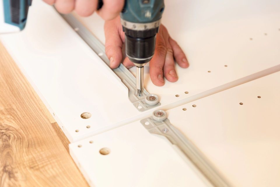 Mani che usano un trapano per installare la ferramenta su un pannello bianco di un armadio