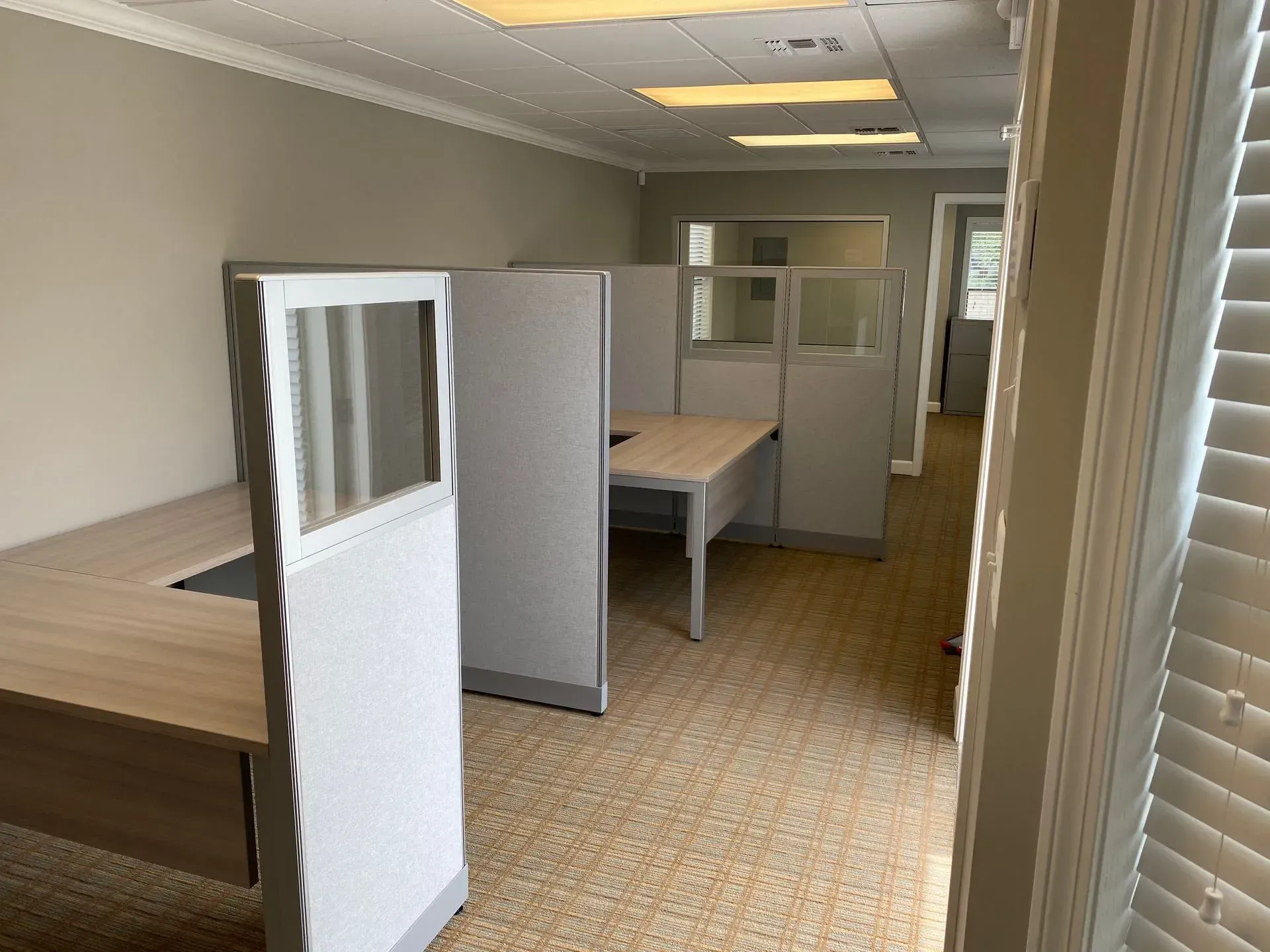 Office cubicles in a room with neutral-colored walls and a light-colored carpet. Desks are empty.