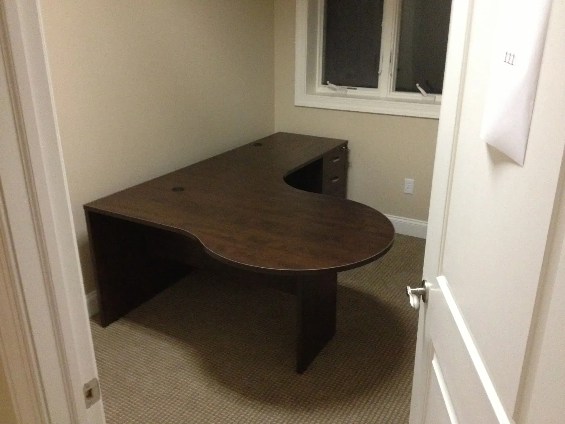 Dark wood L-shaped desk in a small room with beige walls and carpet, near a window and doorway.