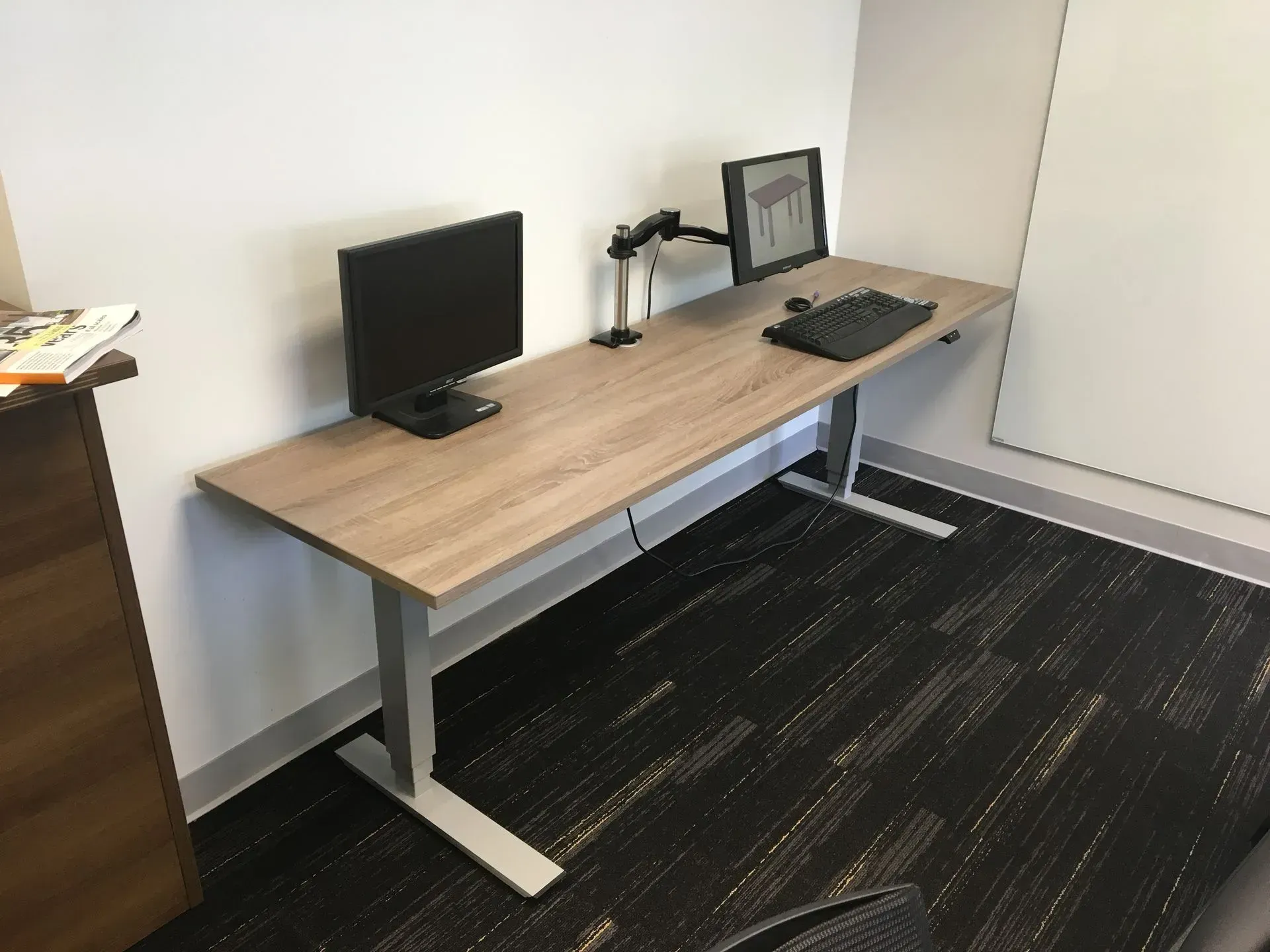 A height-adjustable desk with two monitors, keyboard, and monitor arms against a white wall in an office setting.