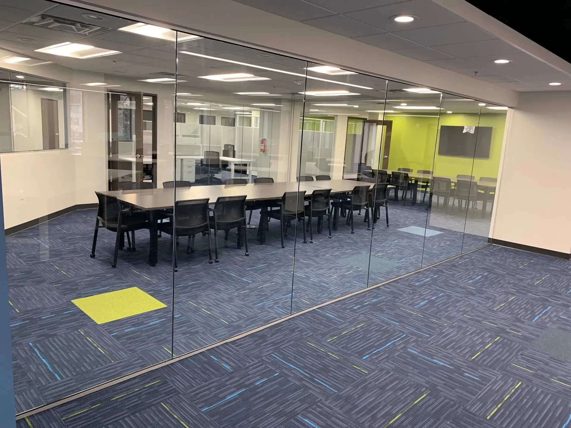 A modern conference room with a long table and chairs, visible through a glass wall, inside a building with blue carpet.