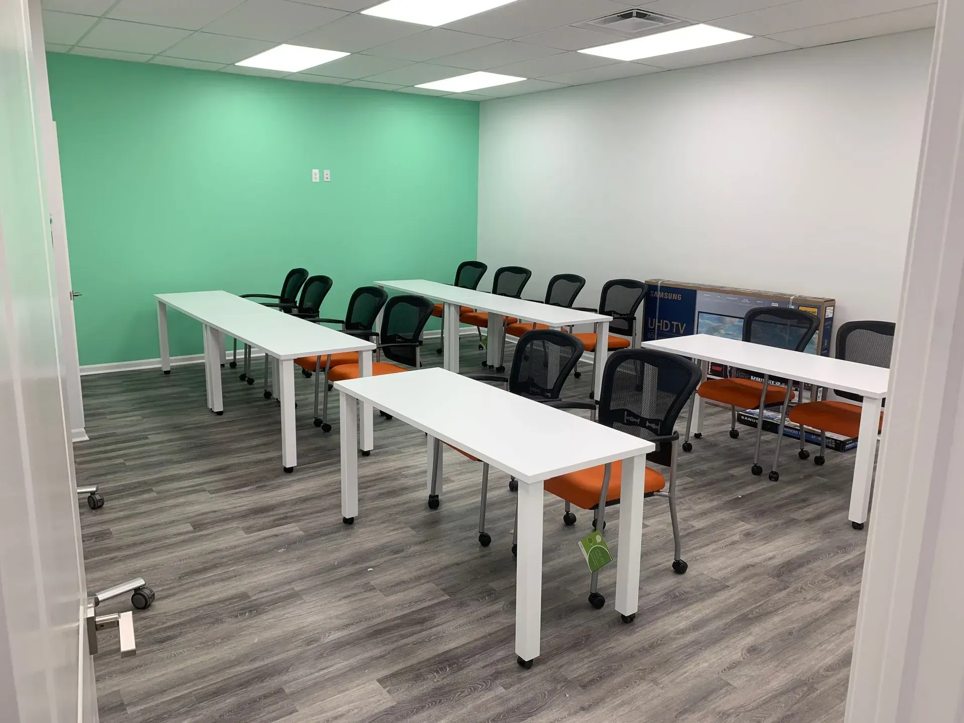 A classroom with white tables and orange chairs against a green and white wall. The door is open.