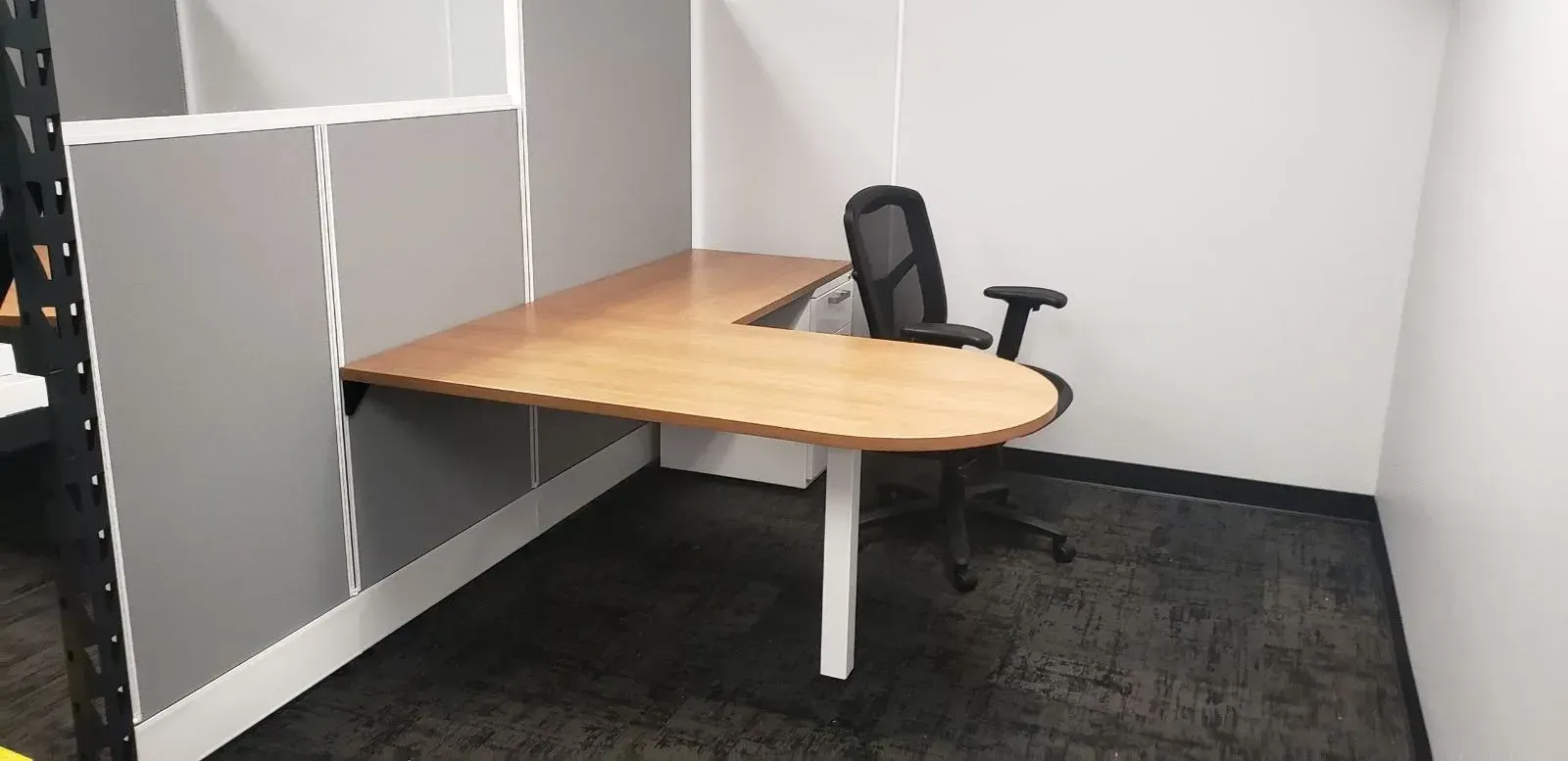 An office cubicle with a light wood-colored desk, black mesh chair, and gray carpet.