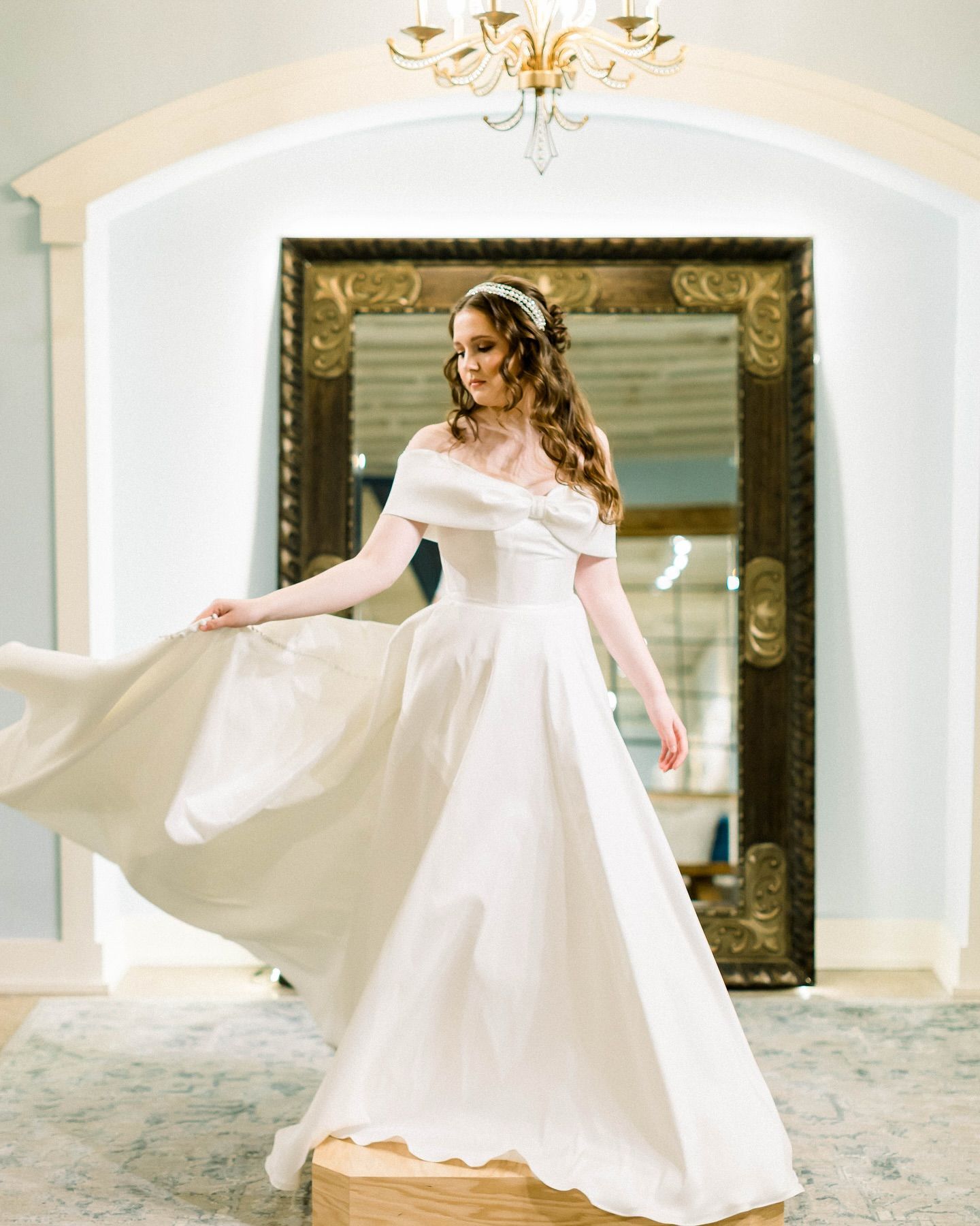 Woman in a white wedding dress twirling with a smile, in front of a large ornate mirror.