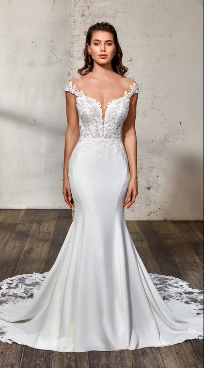 White lace mermaid wedding gown with a long train, shown front view on a model against a gray wall. Domenica -DR2432