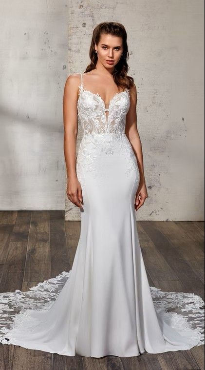Woman in a white lace wedding gown standing against a concrete wall with a long train. Leona - DR2437