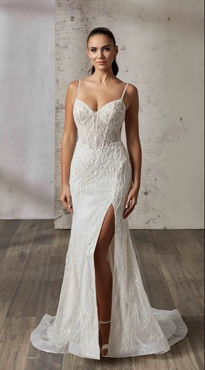 Bridal gown with lace bodice and thigh-high slit, displayed on a model against a concrete wall. Rina - DR2616