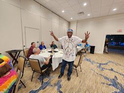 Man posing with peace signs in a banquet room, near tables with seated guests and a doorway in back.