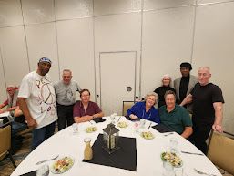 Group of eight people posing around a banquet table in a bright room, with plates and centerpieces.