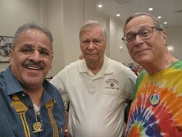 Three smiling men posing indoors, one in a tie-dye shirt and two in collared shirts.