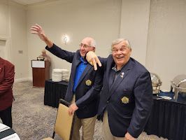 Two men in navy blazers smiling and waving in a conference room
