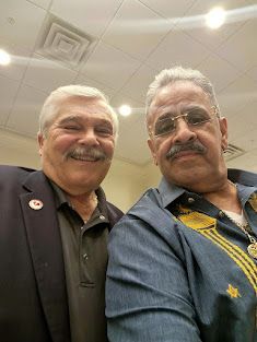 Two men smiling in a selfie indoors, one in a dark jacket and one in a blue shirt with gold embroidery.