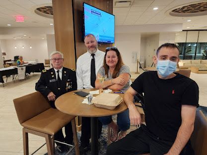 Four people seated at a table in a bright lounge, with a TV screen and a masked man in the foreground.