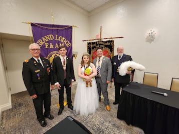 Wedding group posing indoors with purple Grand Lodge banner; bride holds bouquet beside groom and attendants