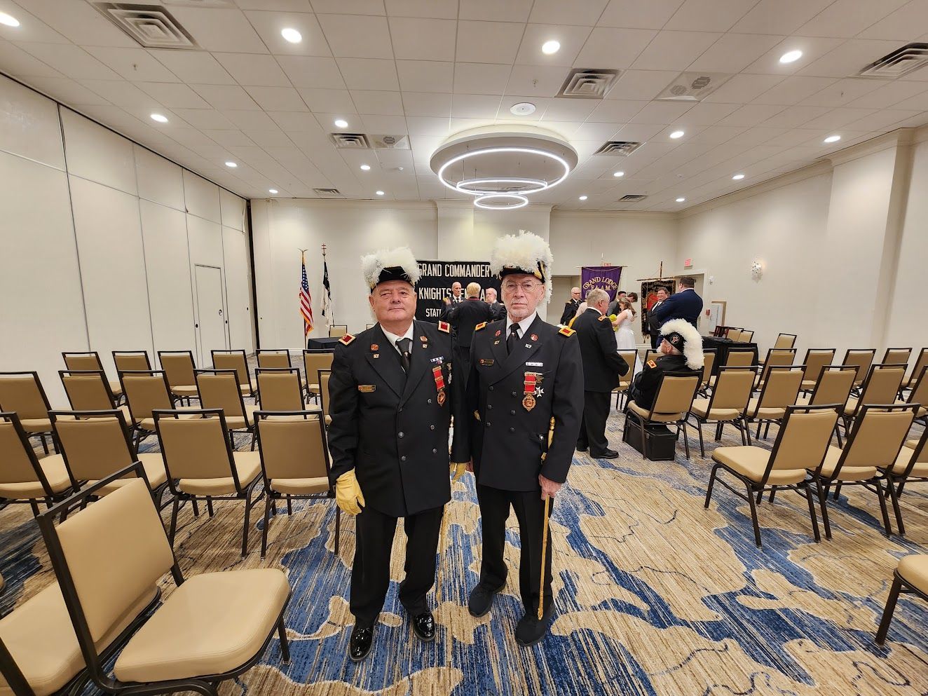 Two uniformed officers stand in an empty banquet hall with rows of chairs and ceiling lights.