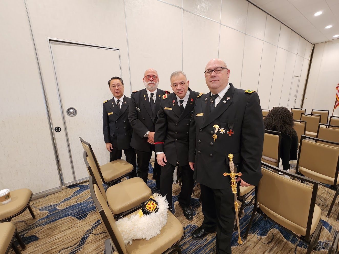 Four men in formal uniforms standing in a conference room beside rows of chairs and a wreath.
