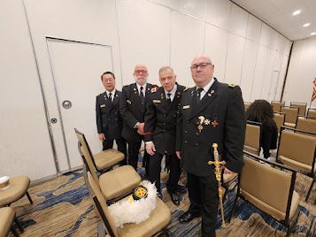 Four men in dark uniforms stand in a conference room beside rows of chairs and a floral wreath.