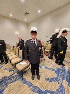 Man in a black uniform with white plumed hat standing in a conference room beside rows of chairs