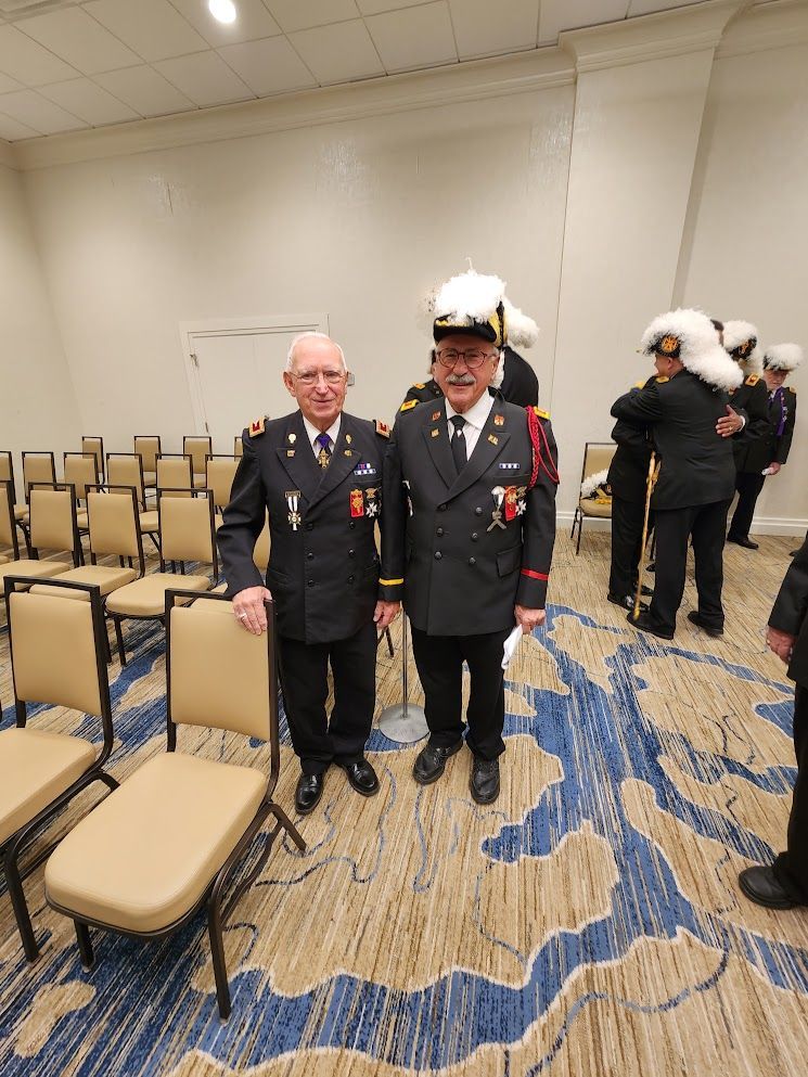Two veterans in dark uniforms stand in a banquet room beside rows of empty chairs.