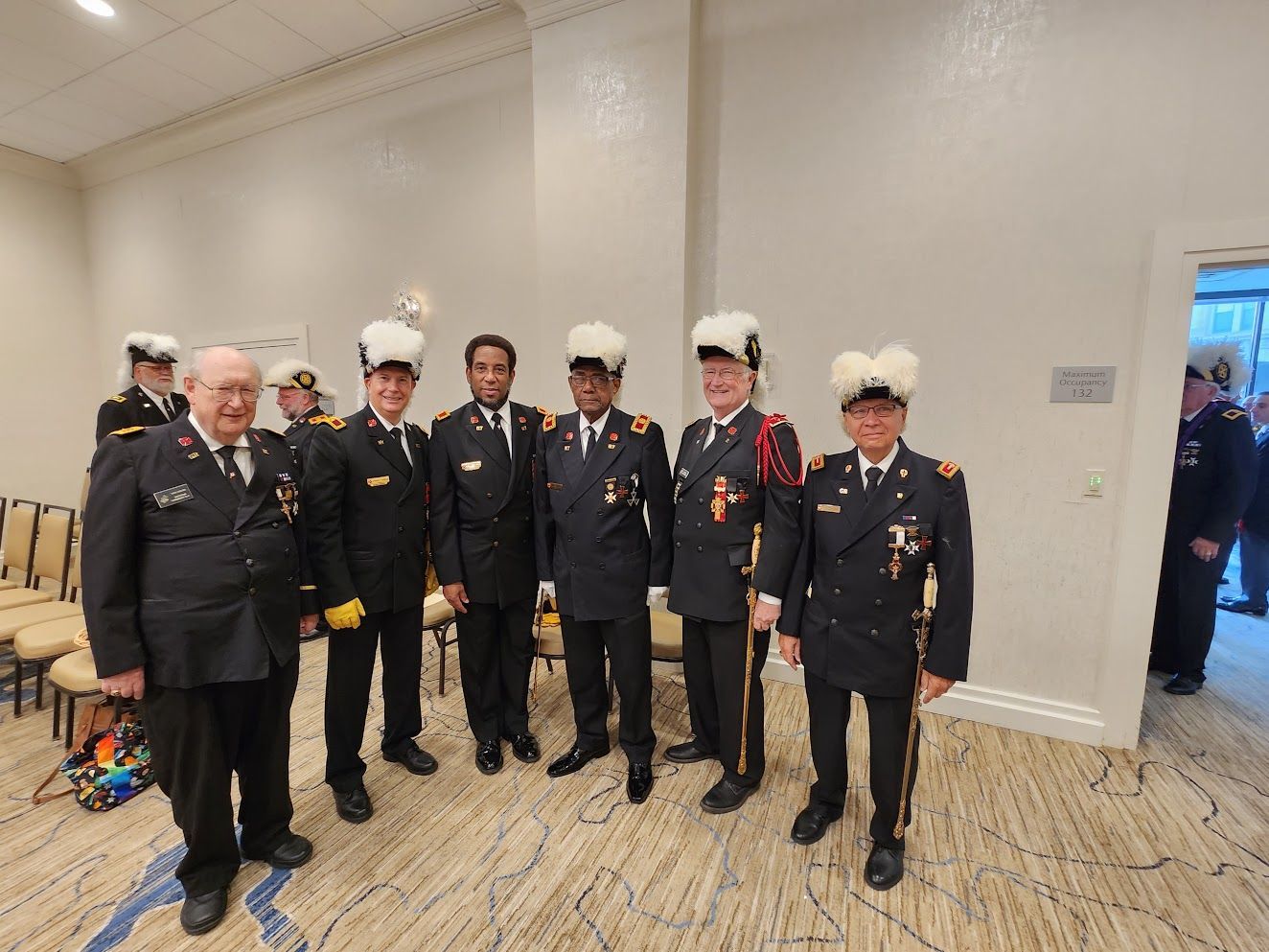 Group of uniformed officers posing indoors at a formal event, with one man in a decorated black suit.