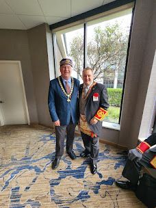 Two people posing indoors by a large window, one in a blue suit and hat, the other in a dark suit with a red armband.