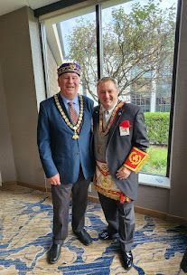 Two people in ceremonial attire stand smiling by a window, wearing medallions and red rosettes.