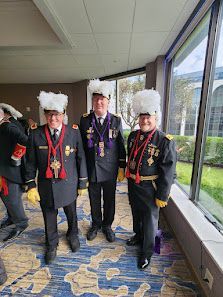 Three people in ceremonial uniforms with fur hats standing in a bright hallway with large windows.