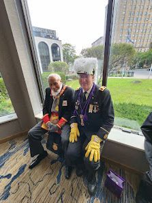 Two seated veterans in dark uniforms with medals, wearing gloves and a white furry hat, by a window overlooking a lawn