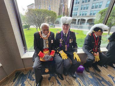 Three people in formal uniforms seated indoors by a window, wearing medals and white hats.