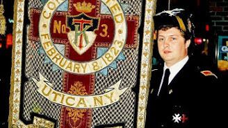 Man in a black suit standing beside a large circular emblem reading 