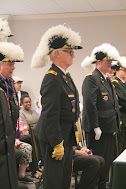 Uniformed officials wearing white plumed hats stand in a line during a ceremony indoors.