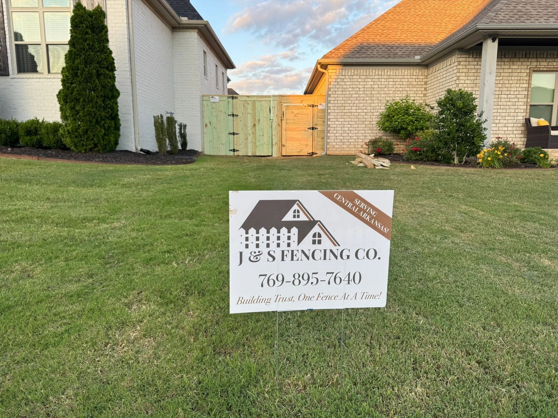 J&S Fence sign on a lawn in front of suburban houses, with a wooden gate in the background