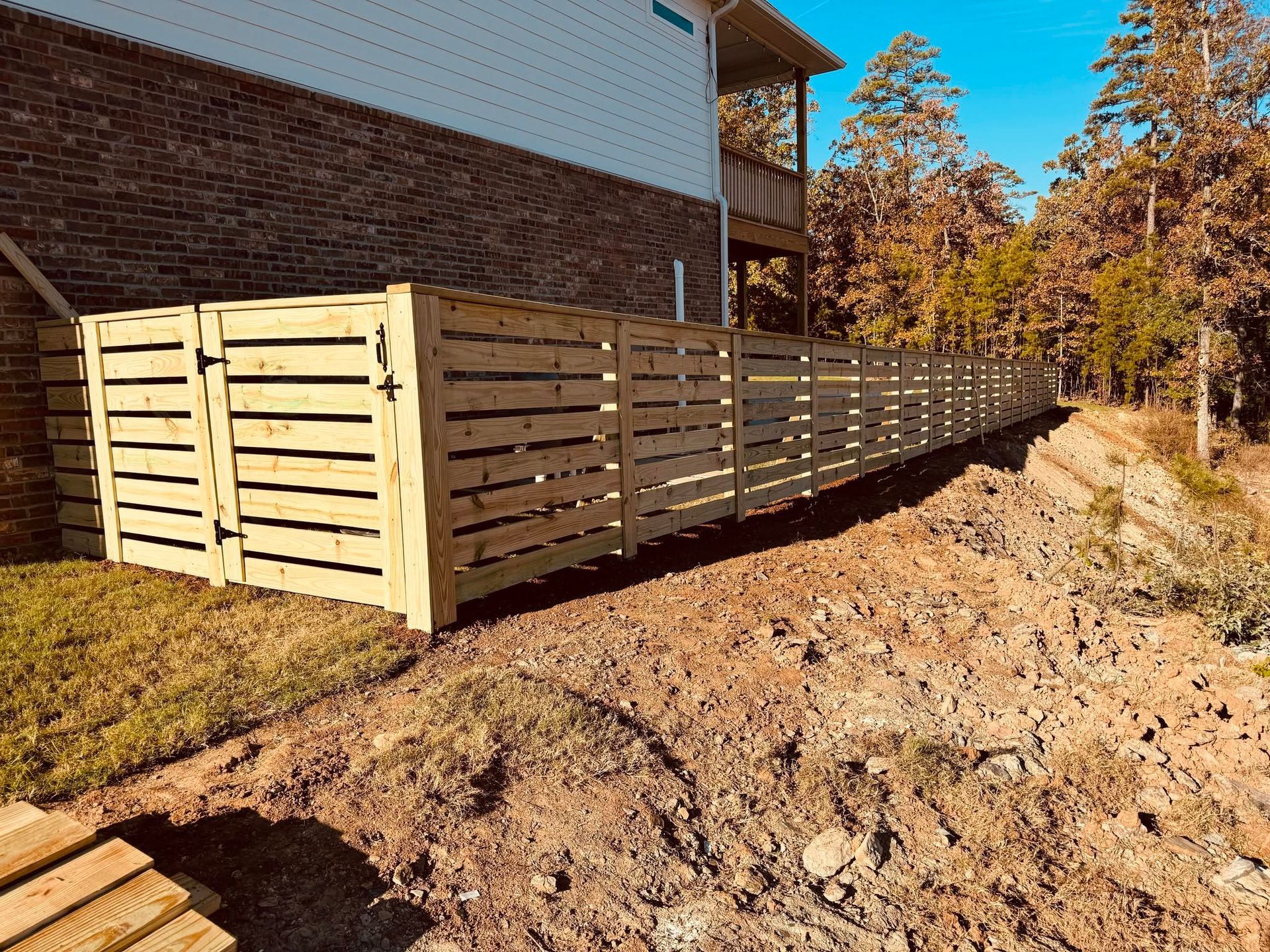 Wooden slat fence beside a brick house on a sloped dirt embankment in autumn