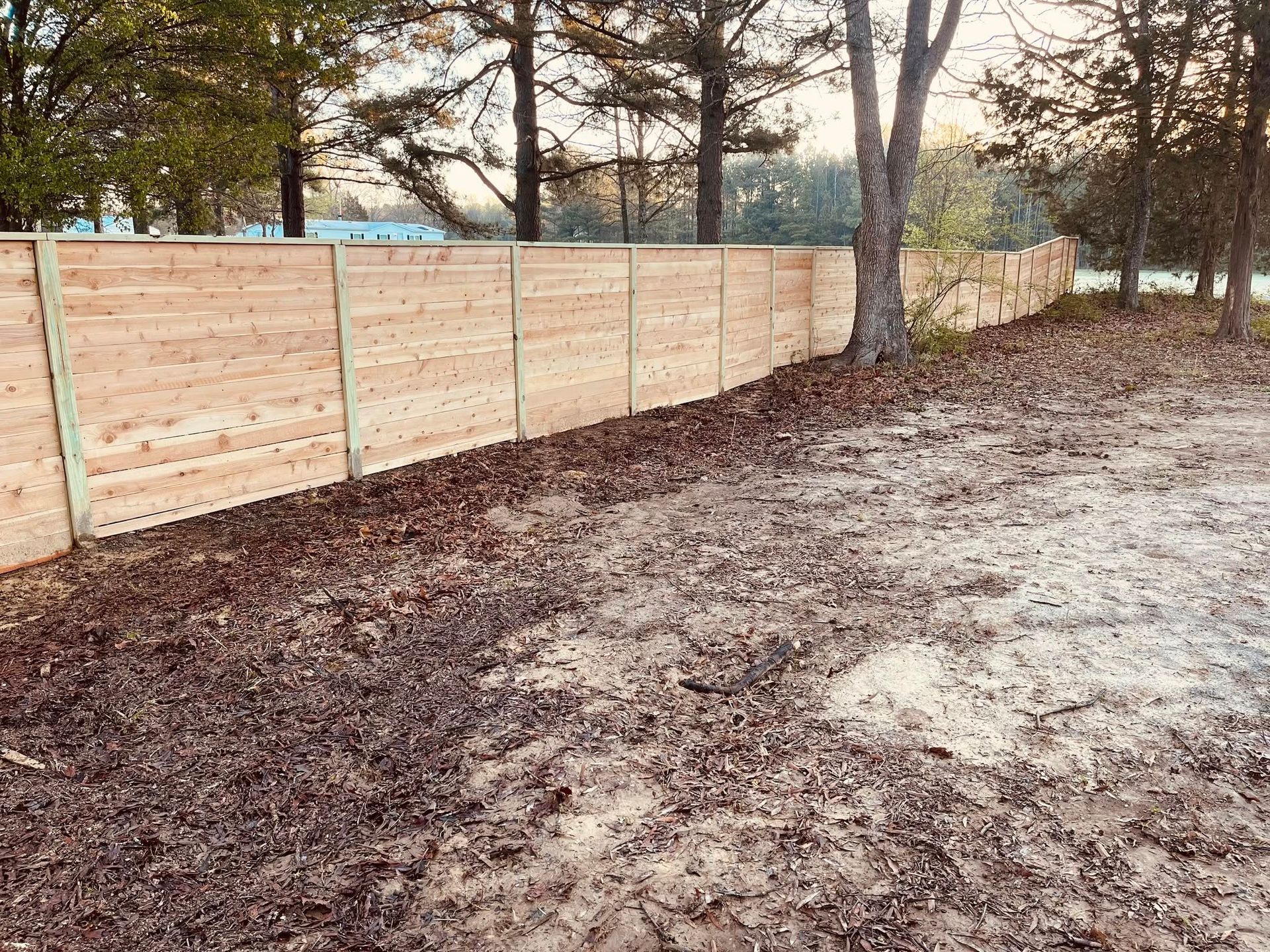 Wooden privacy fence along a wooded yard with bare soil and scattered trees.