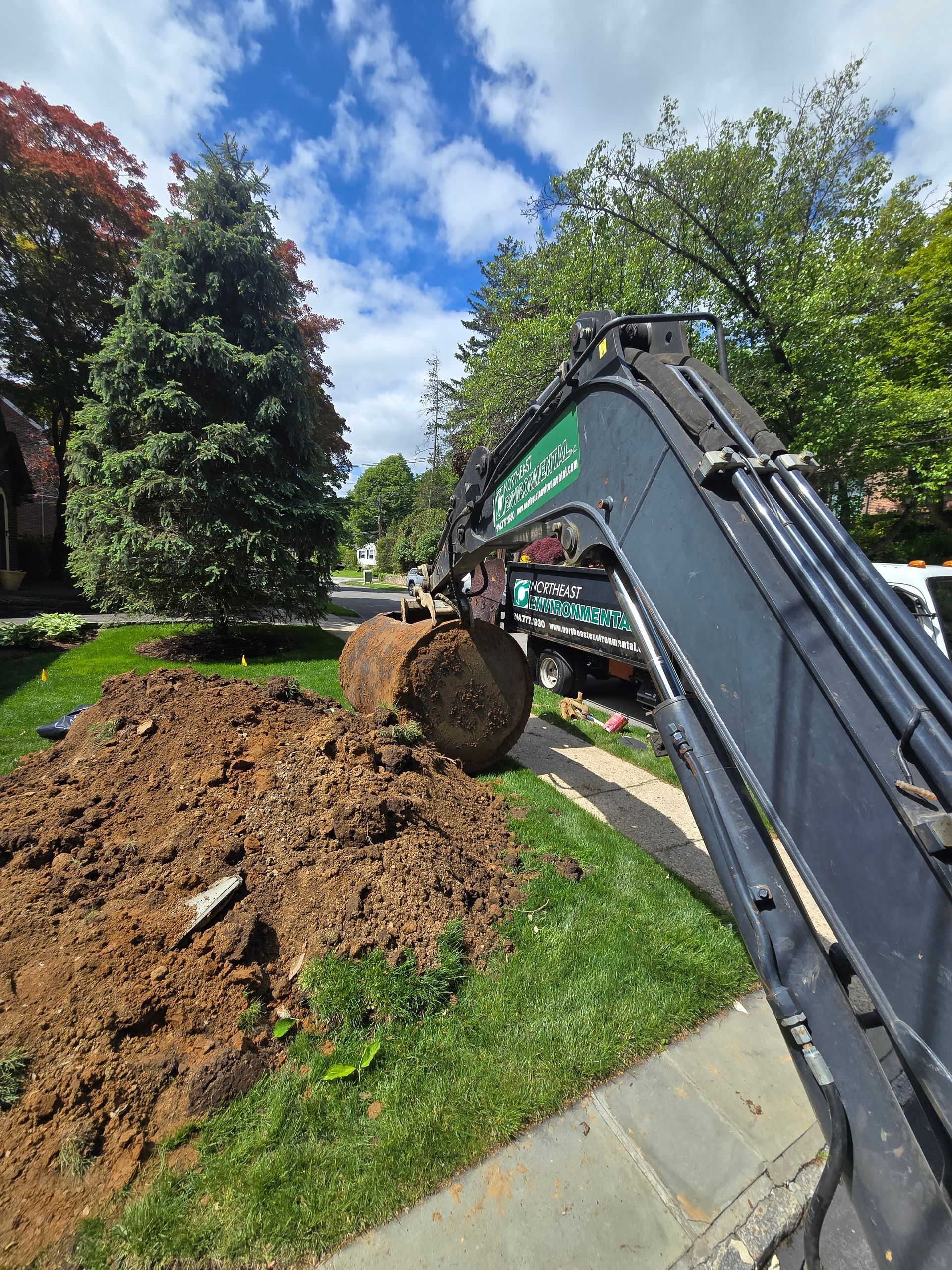 Tank Installation — Westchester County, PA — Northeast Environmental Excavator arm lifting a dirt clump beside a landscaped lawn under a blue sky