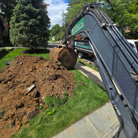 Tank Installation — Westchester County, PA — Northeast Environmental Excavator arm lifting a dirt clump beside a landscaped lawn under a blue sky