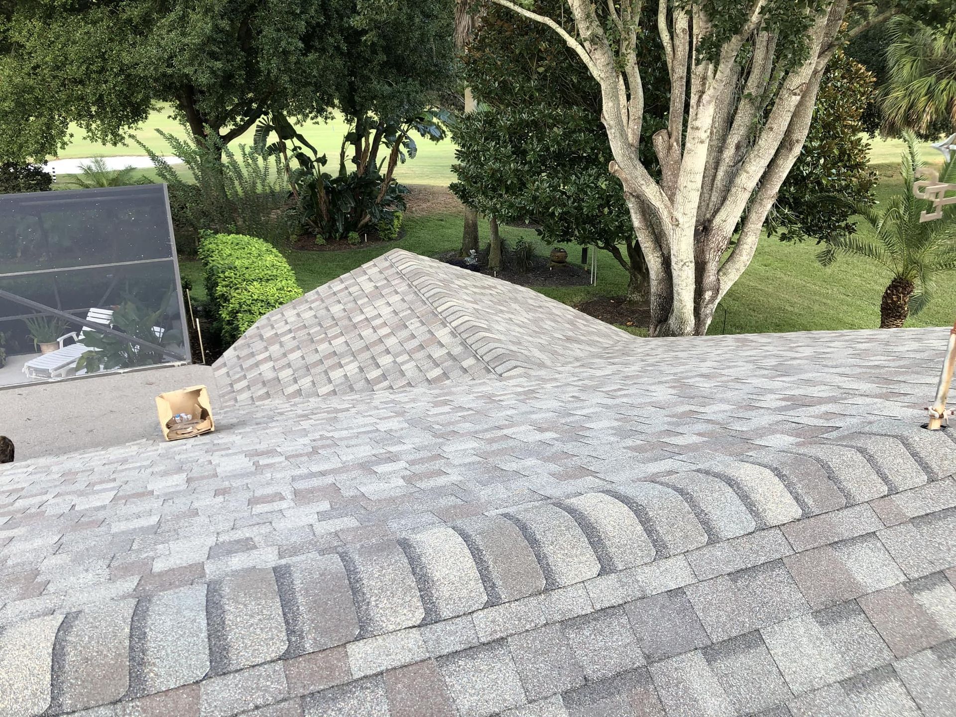 Gray shingle roof with brown and tan shingles. Trees and green lawn in background.