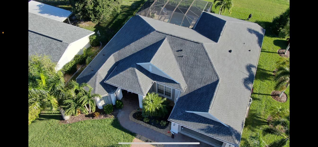 Aerial view of a house with a complex gray roof and green lawn surrounded by trees and a driveway.