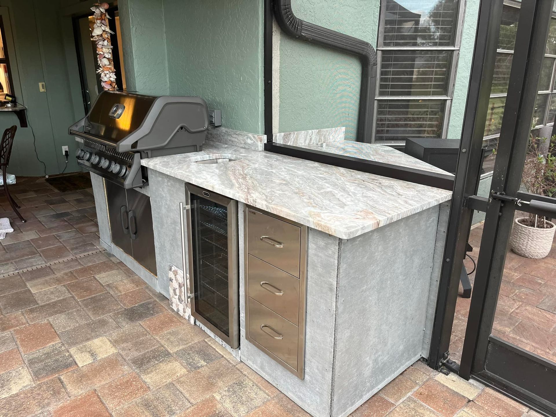 Outdoor kitchen with grill, mini-fridge, drawers, and granite countertop on a brick patio.