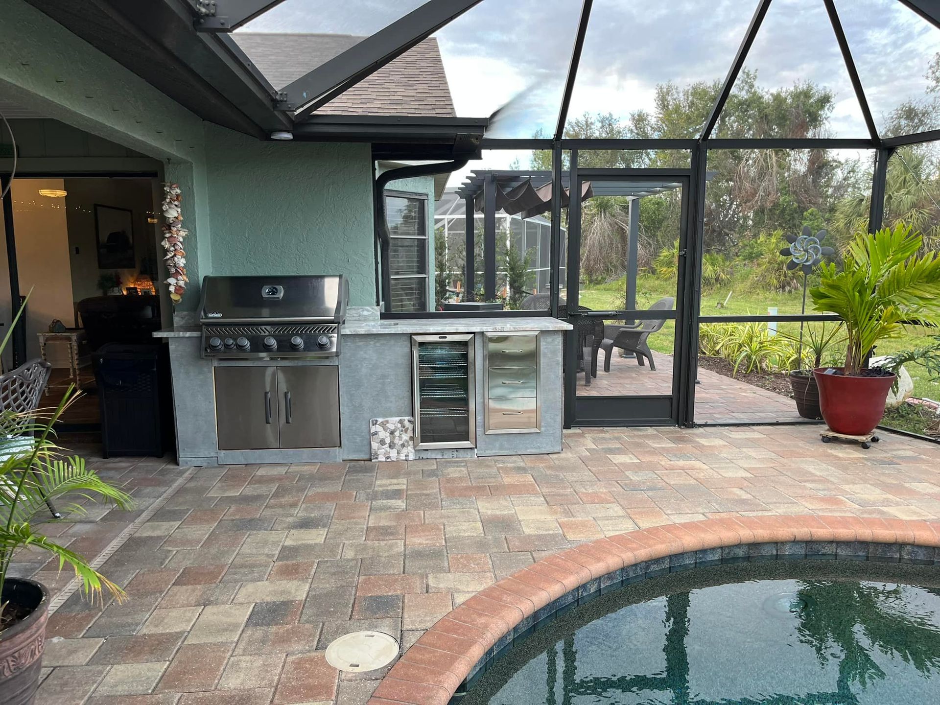Outdoor kitchen with grill, cooler, and pool. Brick patio, screened enclosure, plants.