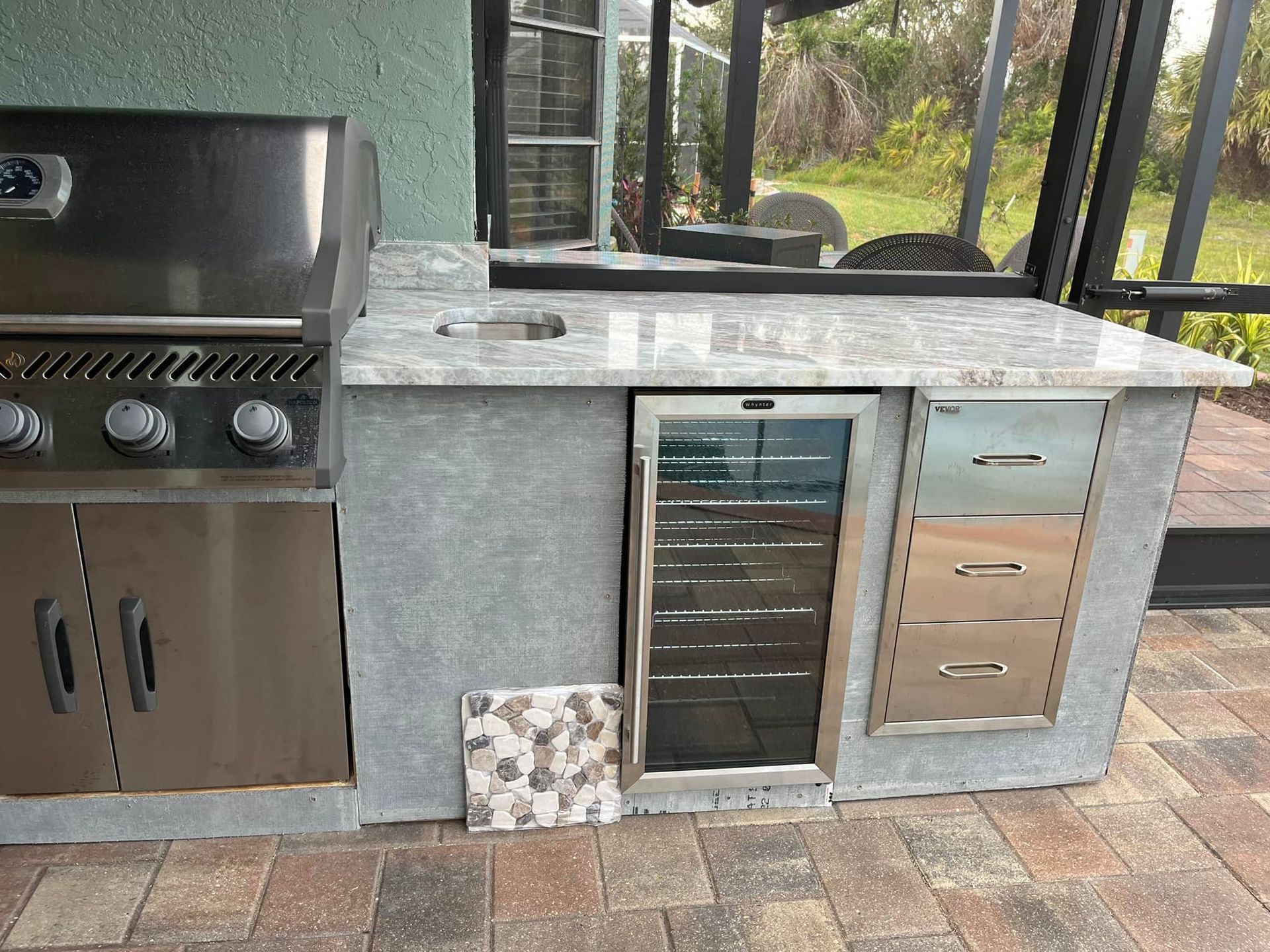 Outdoor kitchen with grill, sink, refrigerator, and drawers on a patio.