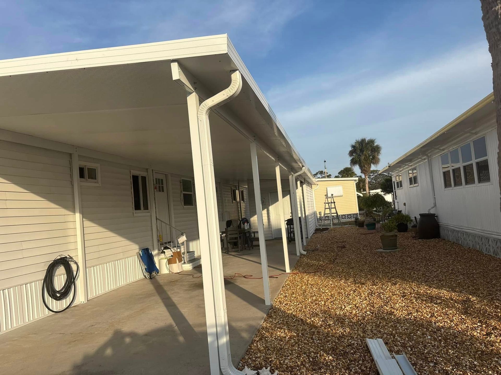 White mobile home with carport, gravel yard, blue sky.