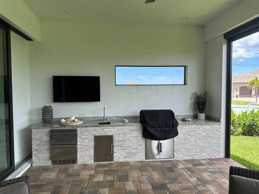Outdoor kitchen with a grill, sink, cabinets, and a TV on a patio.
