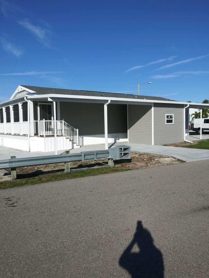 Gray mobile home with porch and steps, blue sky. Shadow of person taking photo.