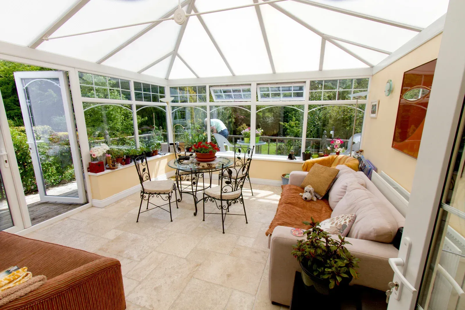 Sunroom with glass walls and roof, furnished with a table, chairs, and sofa, overlooking a garden.